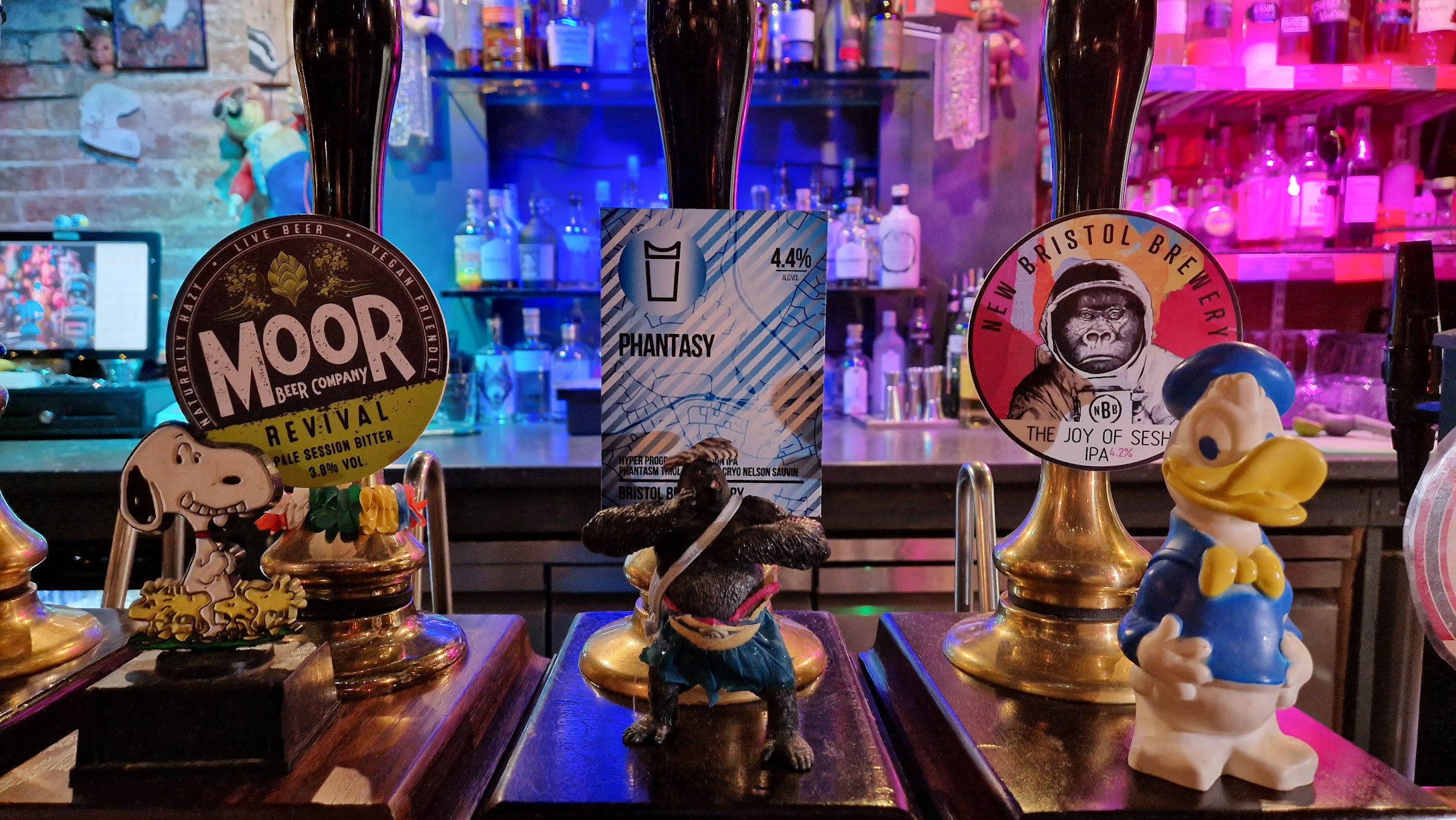 Three beer pumps at a bar, each with a different label of fantastical ales. In front of the pumps are Snoopy, a gorilla figurine, and Donald Duck figures. Colorful bottles are visible on shelves in the background.