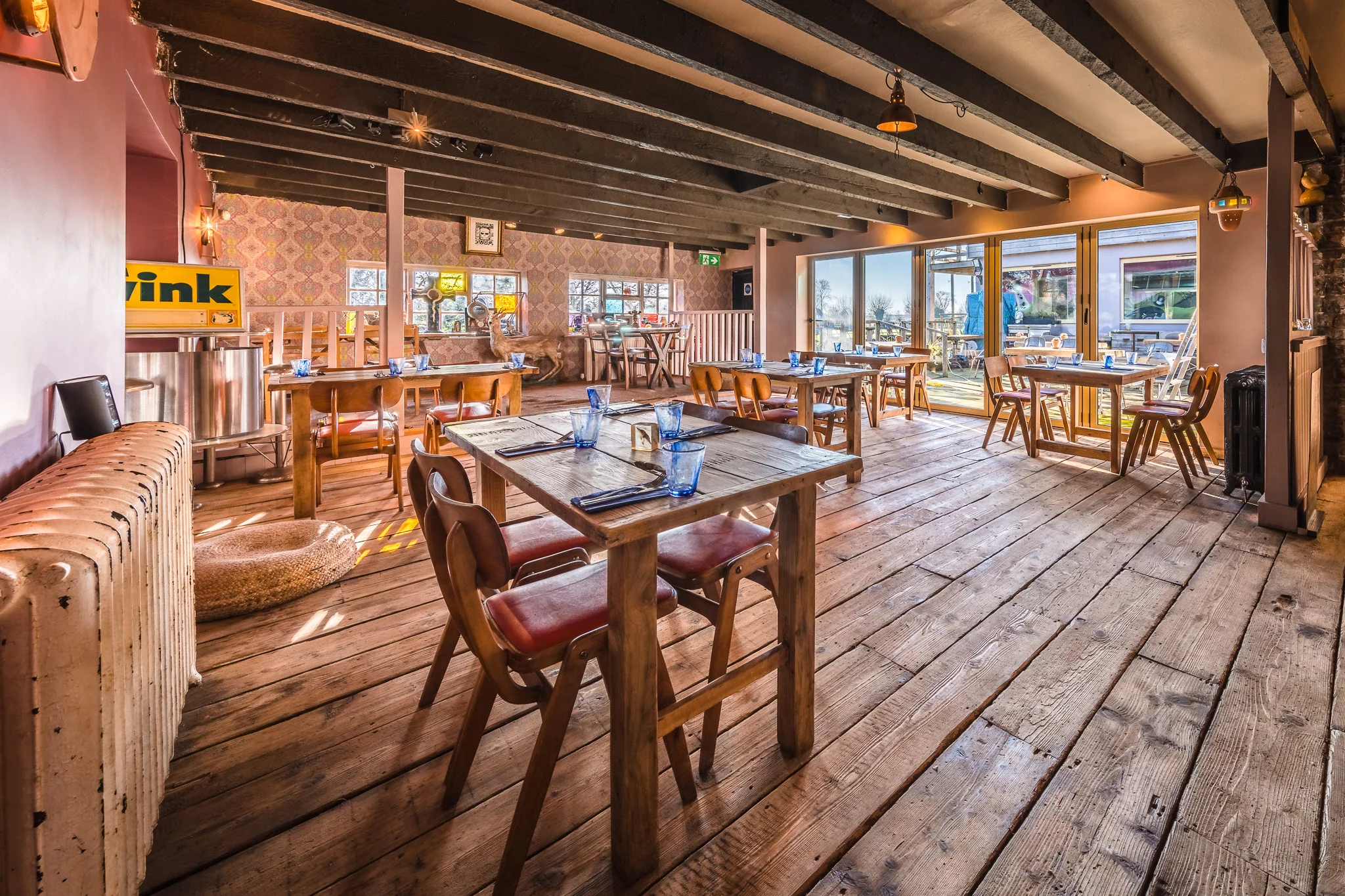 A cozy, sunlit restaurant dining area with wooden floors, tables set with blue glasses and cutlery, leather chairs, and large windows letting in natural light. Warm, rustic decor and exposed ceiling beams create an inviting atmosphere.