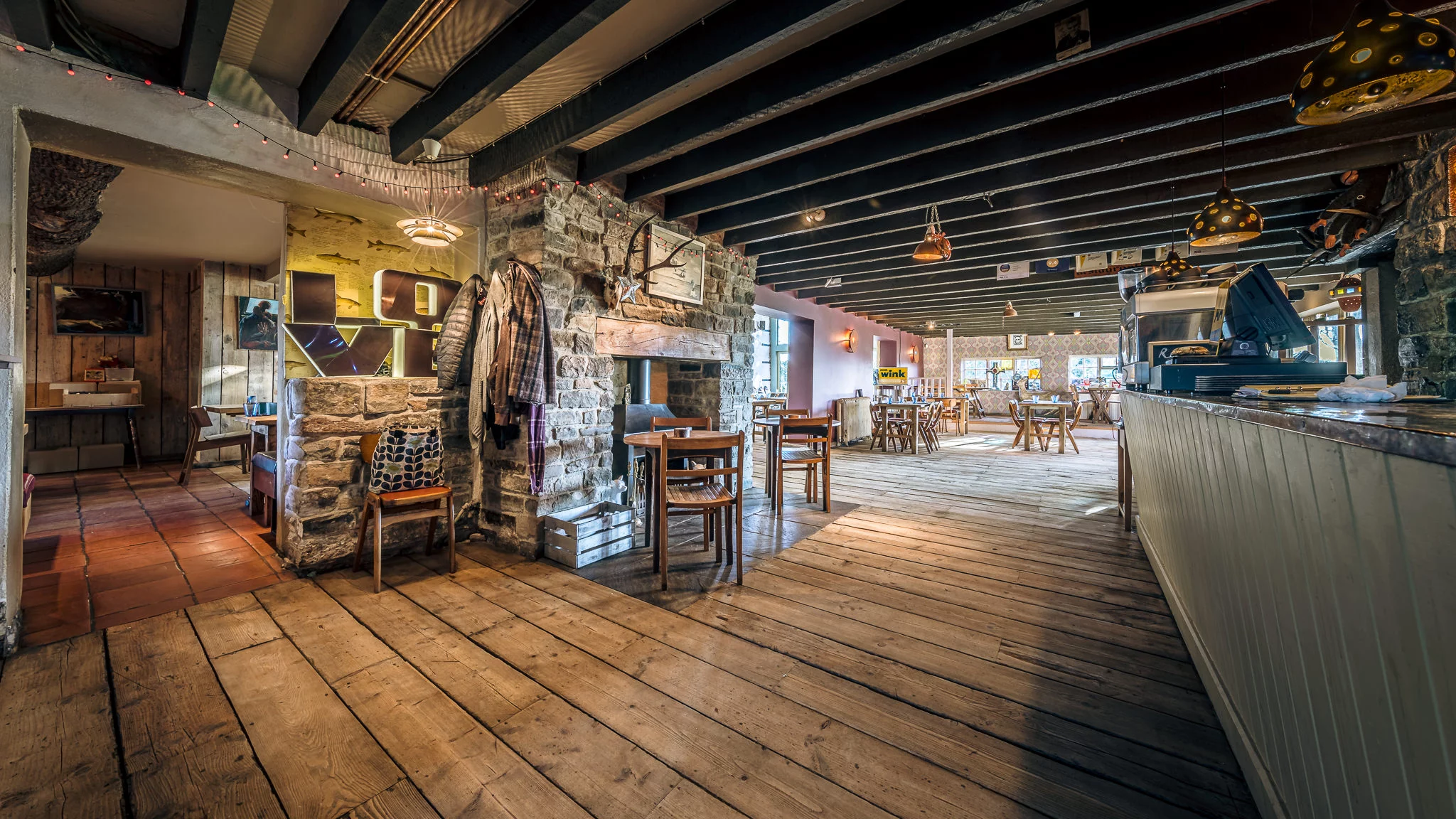 A cozy café interior with stone walls, wooden floors, scattered tables and chairs, warm lighting, and rustic decor. A coat hangs by a stone fireplace, and sunlight streams in from large windows at the far end.