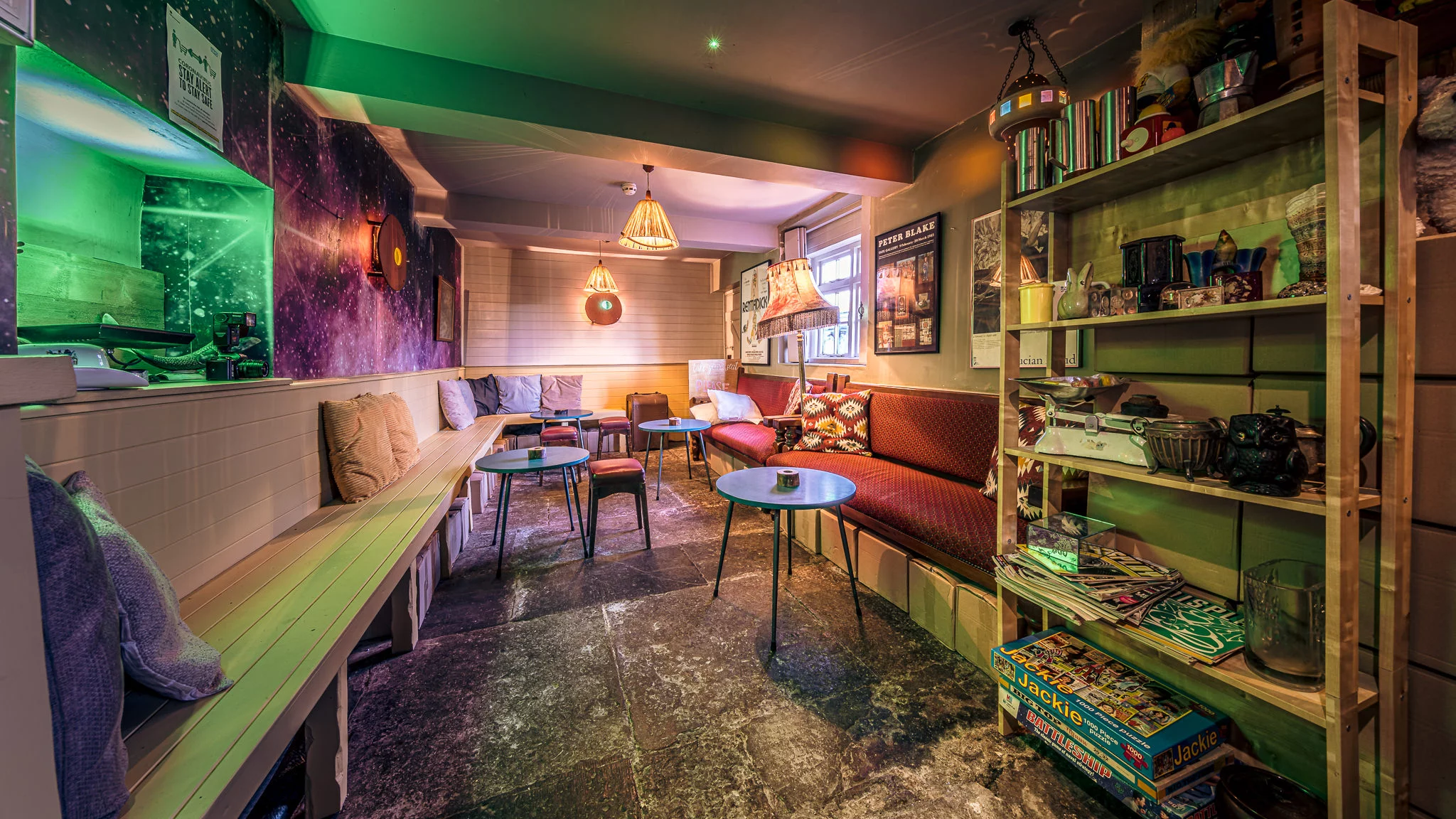 Cozy cafe with colorful lighting, cushioned benches, small round tables, and shelves holding board games, books, and decor items. Warm and inviting atmosphere with art on the walls and a relaxed seating arrangement.