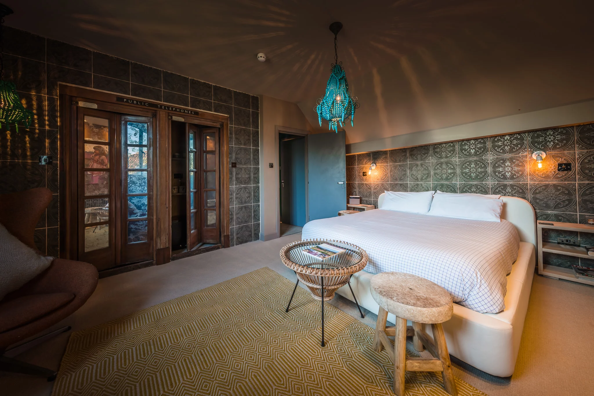 A cozy bedroom with a large bed, patterned wall tiles, two turquoise hanging lights, a wicker table and stool, and an armchair. Wooden doors with glass panels lead to another room. The space feels warm and artistic.
