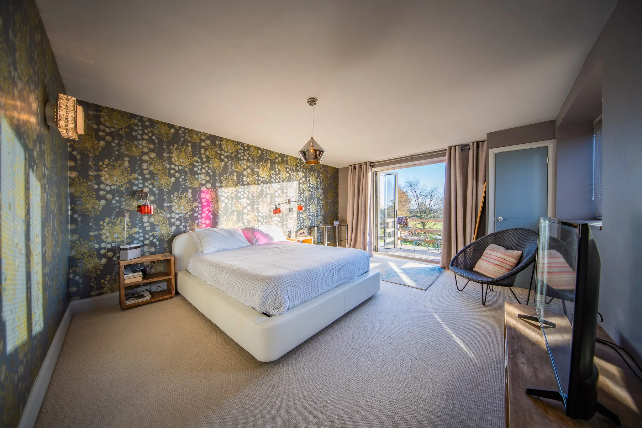 Bright bedroom with patterned wallpaper, a white bed, bedside tables, modern lighting, TV, chairs, and large glass doors opening to a balcony with a scenic outdoor view. Sunlight fills the room.