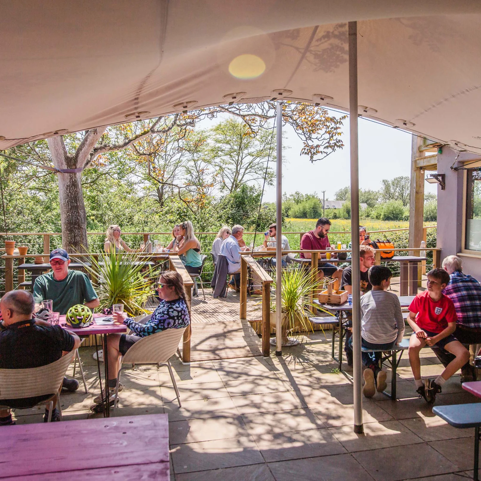 People sit at outdoor tables under a canopy, enjoying food and drinks in a sunny, leafy garden setting. Some chat, while others relax and look at the view. Lush greenery and trees are visible in the background.