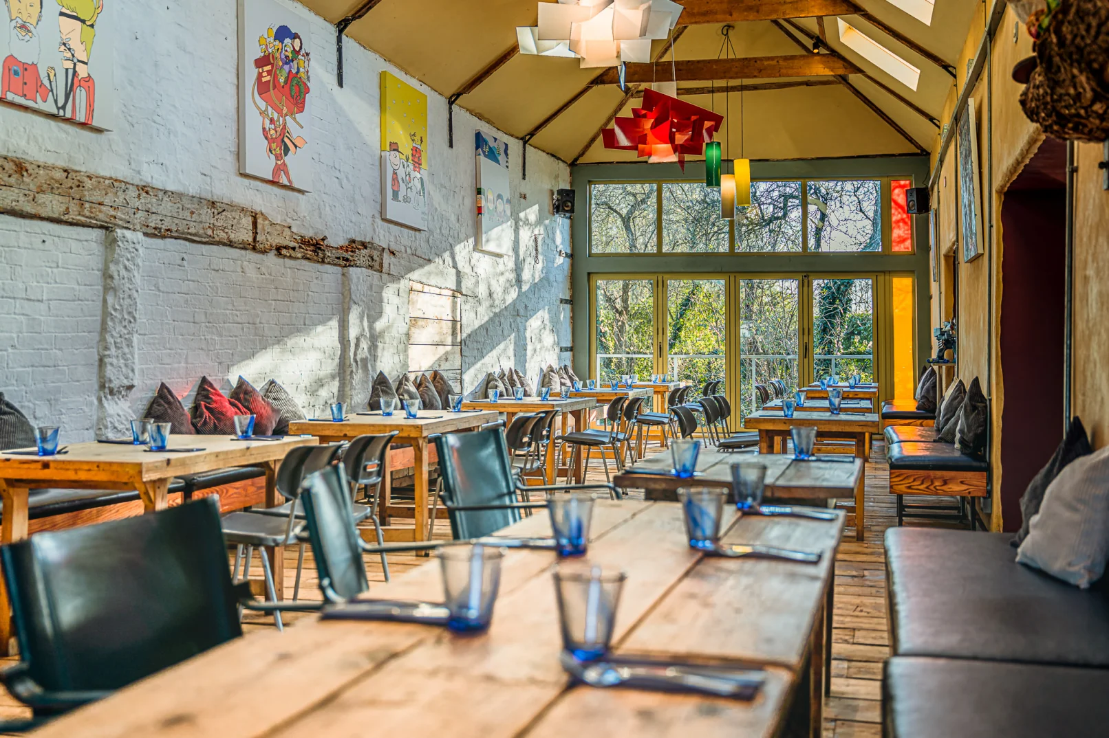 A bright, modern restaurant with wooden tables, black chairs, and blue glasses, sunlight streaming through large windows, colorful artwork on white brick walls, and cushions on benches along the sides.