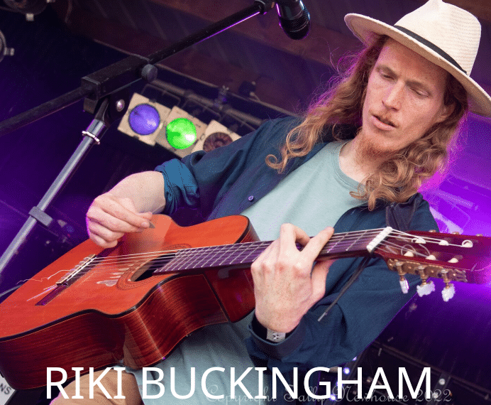 A man with long hair and a hat plays an acoustic guitar on stage under colorful lights. Ricki Buckingham’s name is boldly displayed in large white letters at the bottom of the image.